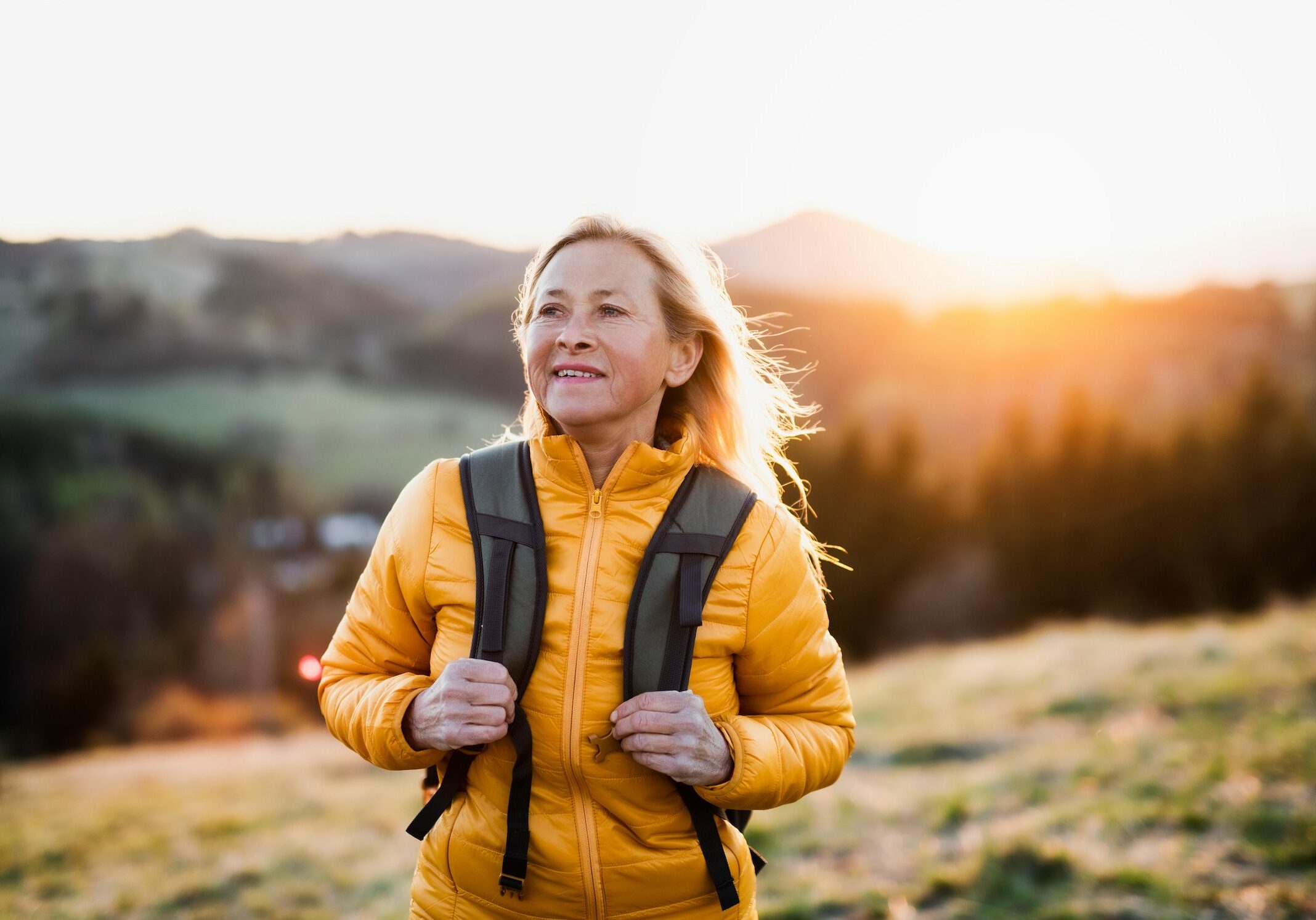 Attractive senior woman with backpack walking outdoors in nature at sunset, hiking.